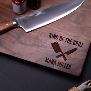 May include: A walnut wood cutting board with the text "King of the Grill" and "Mark Miller" engraved on it. Two crossed butcher knives are also engraved on the board.