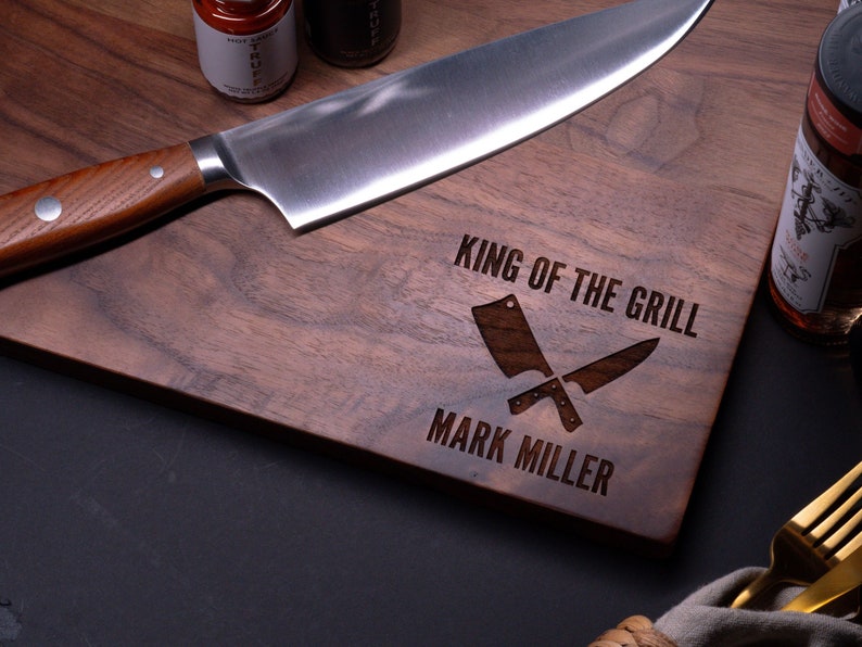 May include: A walnut wood cutting board with the text "King of the Grill" and "Mark Miller" engraved on it. Two crossed butcher knives are also engraved on the board.