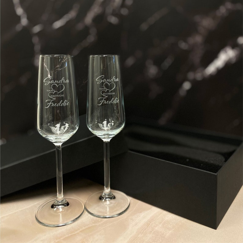 May include: Two clear glass champagne flutes with the names "Sandra" and "Freddie" etched on them. The flutes are in a black box.