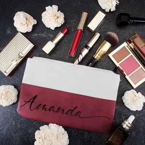 K&ouml;nnte beinhalten: Eine rote und wei&szlig;e Canvas-Kosmetiktasche mit dem Namen "Amanda" in schwarzer Kursivschrift. Die Tasche ist von verschiedenen Kosmetikprodukten umgeben, darunter Lippenstift, Rouge und Foundation.