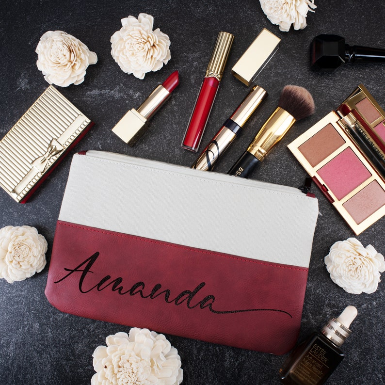 K&ouml;nnte beinhalten: Eine rote und wei&szlig;e Canvas-Kosmetiktasche mit dem Namen "Amanda" in schwarzer Kursivschrift. Die Tasche ist von verschiedenen Kosmetikprodukten umgeben, darunter Lippenstift, Rouge und Foundation.