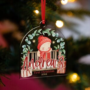 May include: A clear, arch-shaped acrylic Christmas ornament featuring a watercolor illustration of a baby in a red hat and scarf inside a crib. The text reads Andrew's First Christmas 2024, with gift boxes and greenery accents.