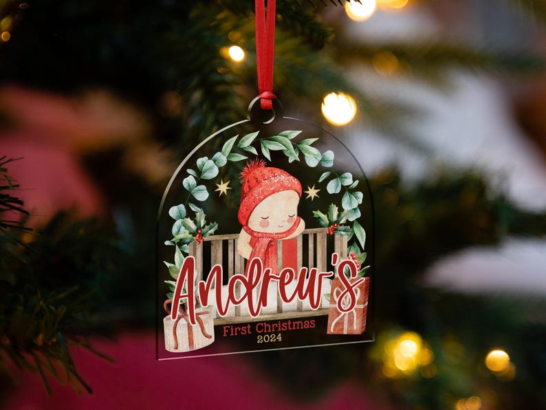 May include: A clear, arch-shaped acrylic Christmas ornament featuring a watercolor illustration of a baby in a red hat and scarf inside a crib. The text reads Andrew's First Christmas 2024, with gift boxes and greenery accents.