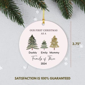 May include: White ceramic Christmas ornament with a pink background. It features three green watercolor trees with the text "Our First Christmas as a Family of Three 2024" and the names "Daddy", "Emily", and "Mommy" underneath the trees. The ornament is 3.75 inches in diameter.