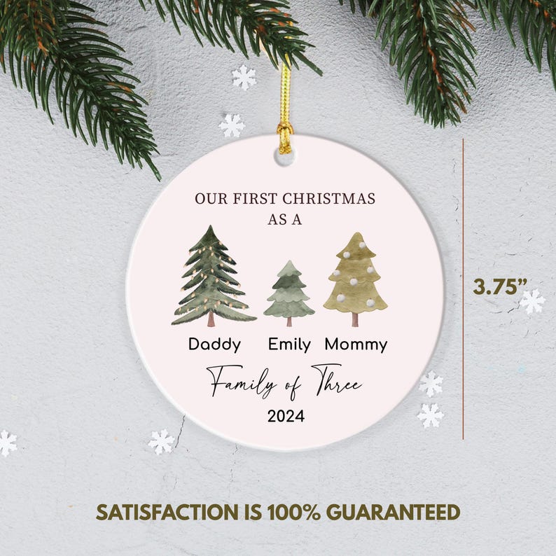 May include: White ceramic Christmas ornament with a pink background. It features three green watercolor trees with the text "Our First Christmas as a Family of Three 2024" and the names "Daddy", "Emily", and "Mommy" underneath the trees. The ornament is 3.75 inches in diameter.