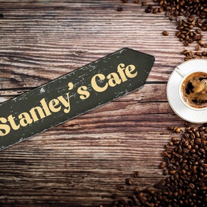 May include: A wooden sign with the text "Stanley's Cafe" in yellow letters on a black background. The sign is on a wooden table with coffee beans scattered around a white cup of coffee.
