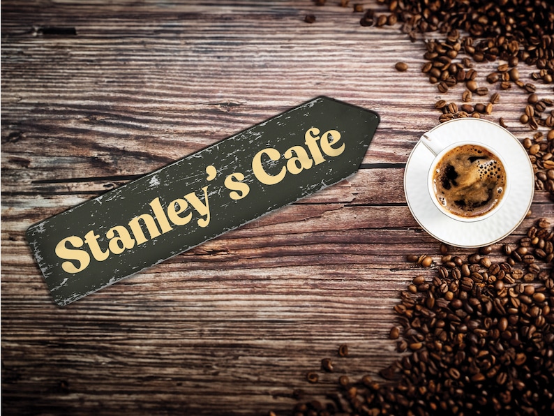May include: A wooden sign with the text "Stanley's Cafe" in yellow letters on a black background. The sign is on a wooden table with coffee beans scattered around a white cup of coffee.