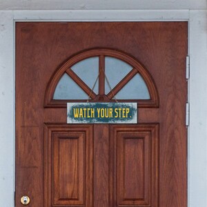 May include: A wooden sign with distressed paint hangs on a brown wooden door. The sign reads "Watch Your Step" in yellow letters.