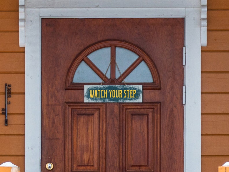 May include: A wooden sign with distressed paint hangs on a brown wooden door. The sign reads "Watch Your Step" in yellow letters.