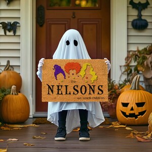 Pu&ograve; includere: Un tappetino da porta marrone con il testo "The Nelsons we smell children" e una grafica di tre streghe con i capelli colorati. Un bambino vestito da fantasma tiene il tappetino.