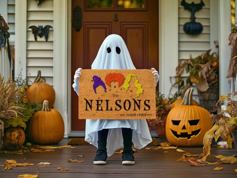 Pu&ograve; includere: Un tappetino da porta marrone con il testo "The Nelsons we smell children" e una grafica di tre streghe con i capelli colorati. Un bambino vestito da fantasma tiene il tappetino.