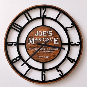 May include: A round wall clock with a wood grain finish and black metal numerals. The center features the text JOE'S MAN CAVE, WHAT HAPPENS IN THE GARAGE STAYS IN THE GARAGE, and EST. 2018. It has black clock hands and a circular frame design.