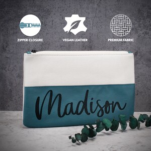 K&ouml;nnte beinhalten: Eine wei&szlig;e und t&uuml;rkisfarbene Rei&szlig;verschlusstasche mit dem Namen "Madison", der in Schwarz auf dem t&uuml;rkisfarbenen Teil gestickt ist.