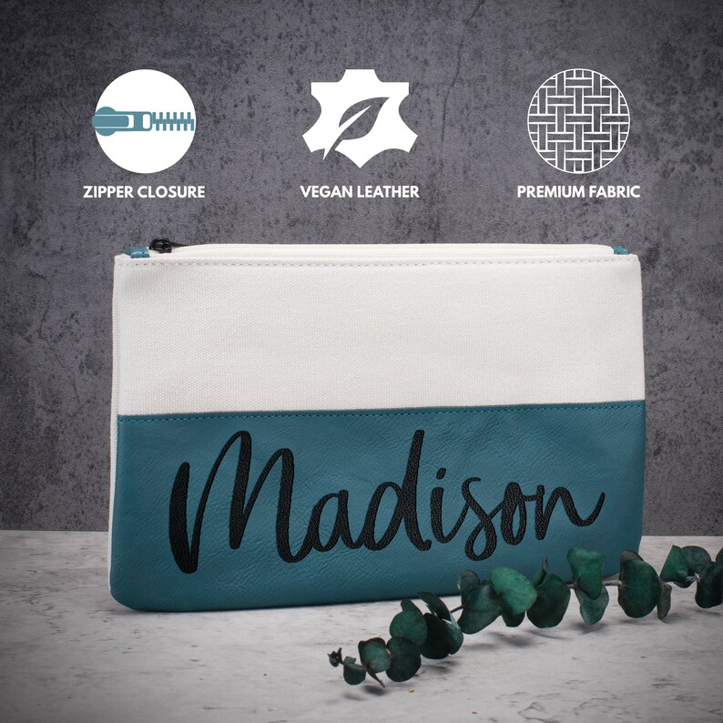 K&ouml;nnte beinhalten: Eine wei&szlig;e und t&uuml;rkisfarbene Rei&szlig;verschlusstasche mit dem Namen "Madison", der in Schwarz auf dem t&uuml;rkisfarbenen Teil gestickt ist.