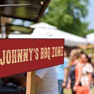 May include: A red wooden sign with white lettering that reads "JOHNNY'S BBQ ZONE". The sign is propped up on a wooden stick and is in front of a blurred background of people and tents.