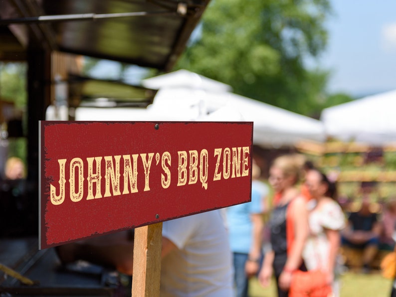 May include: A red wooden sign with white lettering that reads "JOHNNY'S BBQ ZONE". The sign is propped up on a wooden stick and is in front of a blurred background of people and tents.