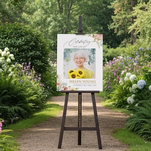 May include: A memorial sign on a wooden easel in a garden setting. The sign reads "Celebrating the Life of Helen Young" with a photo of a woman holding sunflowers. Floral accents frame the photo and text.