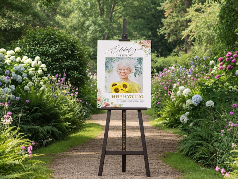 May include: A memorial sign on a wooden easel in a garden setting. The sign reads "Celebrating the Life of Helen Young" with a photo of a woman holding sunflowers. Floral accents frame the photo and text.