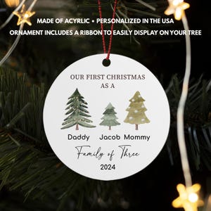 May include: White round Christmas ornament with green watercolor trees and the text "Our First Christmas as a Family of Three 2024" with the names "Daddy", "Jacob", and "Mommy" under each tree.