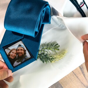 May include: A teal blue tie with a small square photo of a couple in the center. The photo is framed with a black border. The tie is on a white shirt.