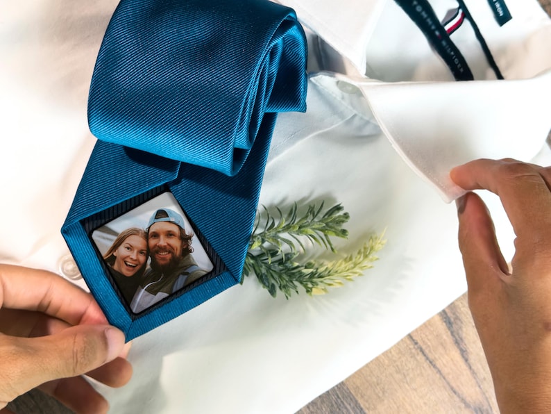 May include: A teal blue tie with a small square photo of a couple in the center. The photo is framed with a black border. The tie is on a white shirt.