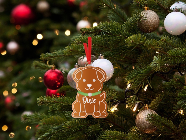 May include: A gingerbread dog ornament with the name "Dixie" on it hangs on a Christmas tree with red and gold ornaments.