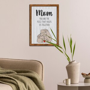May include: A framed print with a white background and wood-grain frame. The print features a drawing of three bears hugging with a heart in the center. The text reads "Mom you are the piece that holds us together".