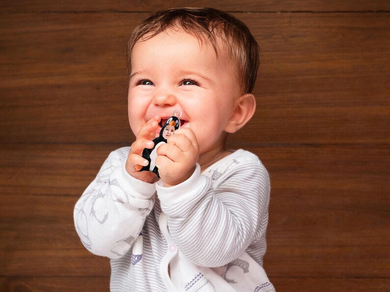 Pu&ograve; includere: Un bambino che indossa un body a righe bianche e grigie sta ridendo mentre tiene in mano un giocattolo di pinguino bianco e nero. Il giocattolo ha una piccola faccia.
