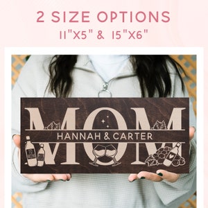 May include: A brown wooden sign with the word "MOM" in large, white letters. The sign also includes the names "HANNAH & CARTER" and images of wine bottles, glasses, and grapes.