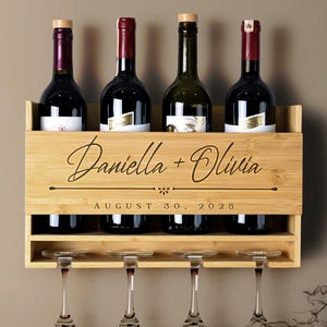 May include: A wooden wine rack with a personalized inscription that reads "Daniella + Olivia August 30, 2025". The rack holds four bottles of wine and has four glass holders underneath.