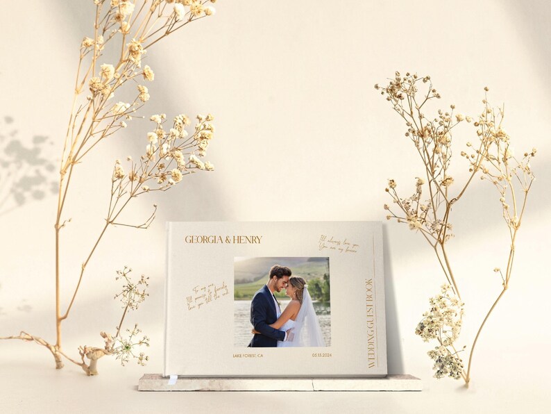 Puede incluir: Un libro de invitados de boda blanco con letras doradas. La portada presenta los nombres "Georgia & Henry" y una foto de una pareja en un campo. El libro tambi&eacute;n incluye la fecha "06.13.2021" y la ubicaci&oacute;n "Lake Tahoe, CA".