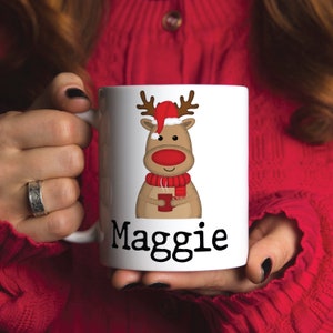 Puede incluir: Taza de cer&aacute;mica blanca con un reno de dibujos animados que lleva un gorro de Pap&aacute; Noel y sostiene una taza roja. La taza est&aacute; personalizada con el nombre "Maggie".
