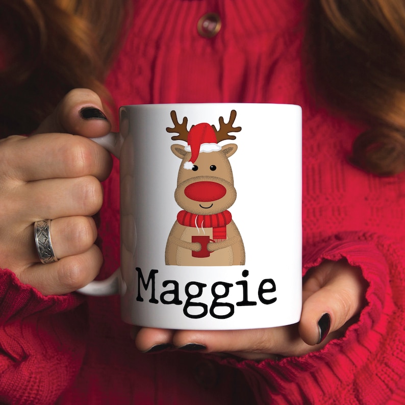 Puede incluir: Taza de cer&aacute;mica blanca con un reno de dibujos animados que lleva un gorro de Pap&aacute; Noel y sostiene una taza roja. La taza est&aacute; personalizada con el nombre "Maggie".