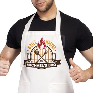 May include: White apron with a brown and orange grill master design. The design includes a grill, crossed grilling tools, flames, and the text "Grill Master" and "Michael's BBQ".