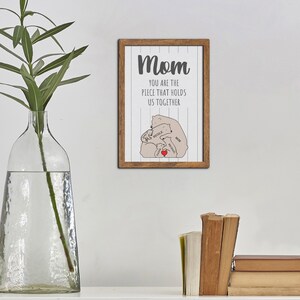 May include: A framed print with a white background and gray text that reads "Mom you are the piece that holds us together". The print features a stylized illustration of a mother bear and three cubs.