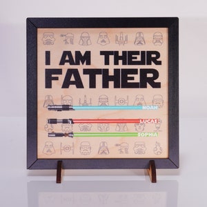 May include: Wooden framed sign with the text "I AM THEIR FATHER" in black. Below are three lightsabers in blue, red, and green, each with a name. The background features Star Wars character illustrations.