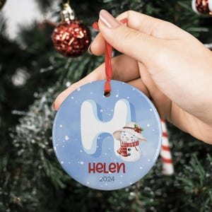 May include: A blue and white ceramic Christmas ornament with a snowman and the letter "H" on it. The ornament also has the name "Helen" and the year "2024" on it.