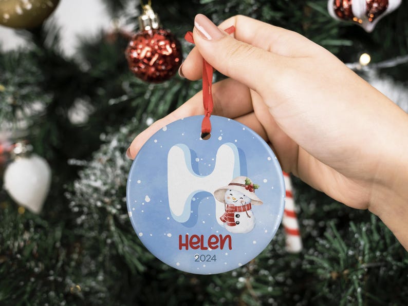 May include: A blue and white ceramic Christmas ornament with a snowman and the letter "H" on it. The ornament also has the name "Helen" and the year "2024" on it.