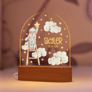 May include: A night light featuring a cartoon bunny on a ladder reaching for a star. The design includes clouds, stars, and the name "Skyler" with the date 08.04.2024. The base is made of wood.