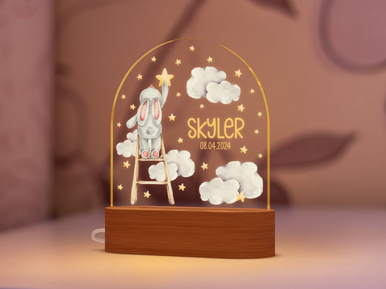 May include: A night light featuring a cartoon bunny on a ladder reaching for a star. The design includes clouds, stars, and the name "Skyler" with the date 08.04.2024. The base is made of wood.