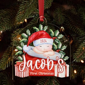 May include: A clear acrylic Christmas ornament featuring a sleeping baby in a red Santa hat with the year 2024, surrounded by holly leaves. The text reads Jacob's First Christmas above two gift boxes with red and white striped wrapping paper.