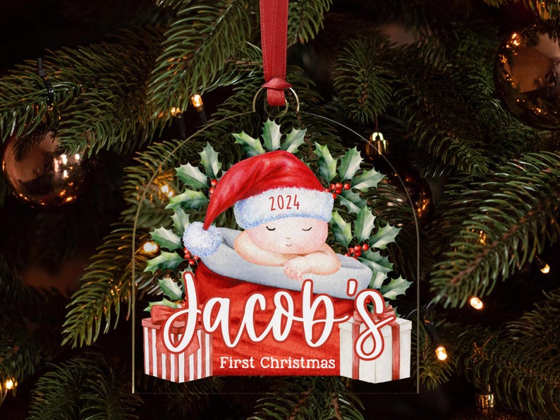 May include: A clear acrylic Christmas ornament featuring a sleeping baby in a red Santa hat with the year 2024, surrounded by holly leaves. The text reads Jacob's First Christmas above two gift boxes with red and white striped wrapping paper.