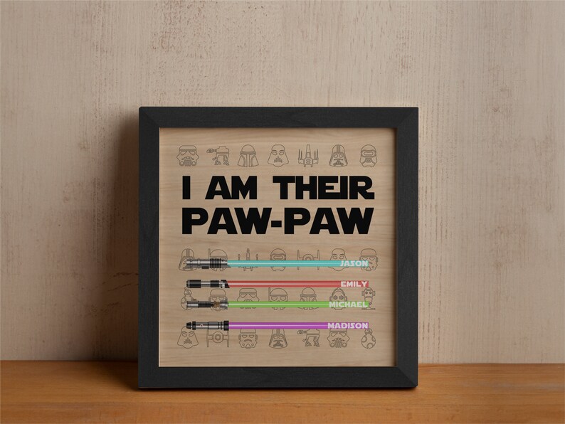 May include: Framed artwork with the text "I AM THEIR PAW-PAW" and lightsaber illustrations. The names Jason, Emily, Michael, and Madison are below the lightsabers. The artwork is in a black frame and set against a wood grain background.