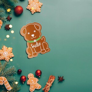 May include: A gingerbread dog ornament with the name "Sadie" on the collar. The ornament is surrounded by Christmas decorations, including a green garland, red ornaments, and gingerbread cookies.