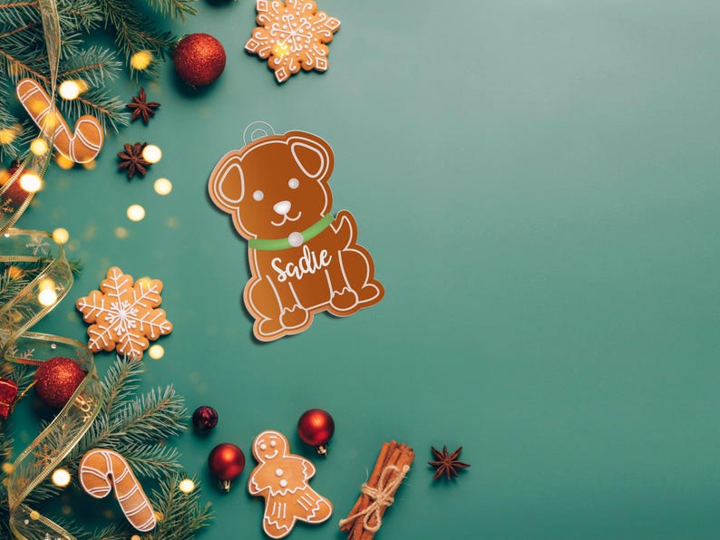 May include: A gingerbread dog ornament with the name "Sadie" on the collar. The ornament is surrounded by Christmas decorations, including a green garland, red ornaments, and gingerbread cookies.