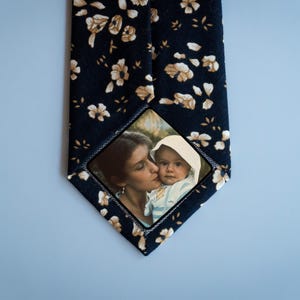 May include: A dark blue tie with a white floral pattern. A small square photo of a woman and a child is sewn onto the tie.