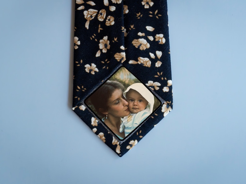 May include: A dark blue tie with a white floral pattern. A small square photo of a woman and a child is sewn onto the tie.