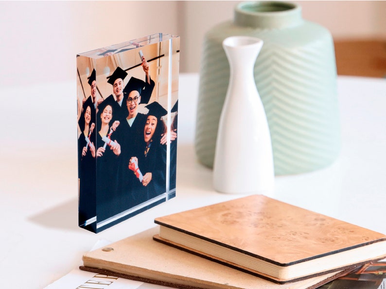 K&ouml;nnte beinhalten: Ein klarer Glasblock mit einem Foto einer Gruppe von Menschen in Abschlusskappen und -gew&auml;ndern, die Diplome halten. Das Foto ist vor einem wei&szlig;en Hintergrund.