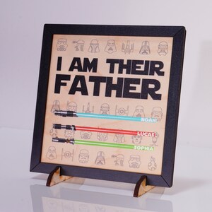 May include: Wooden framed sign with the text "I AM THEIR FATHER" and lightsaber illustrations. The sign includes the names Noah, Lucas, and Sophia. The sign is supported by a wooden stand.
