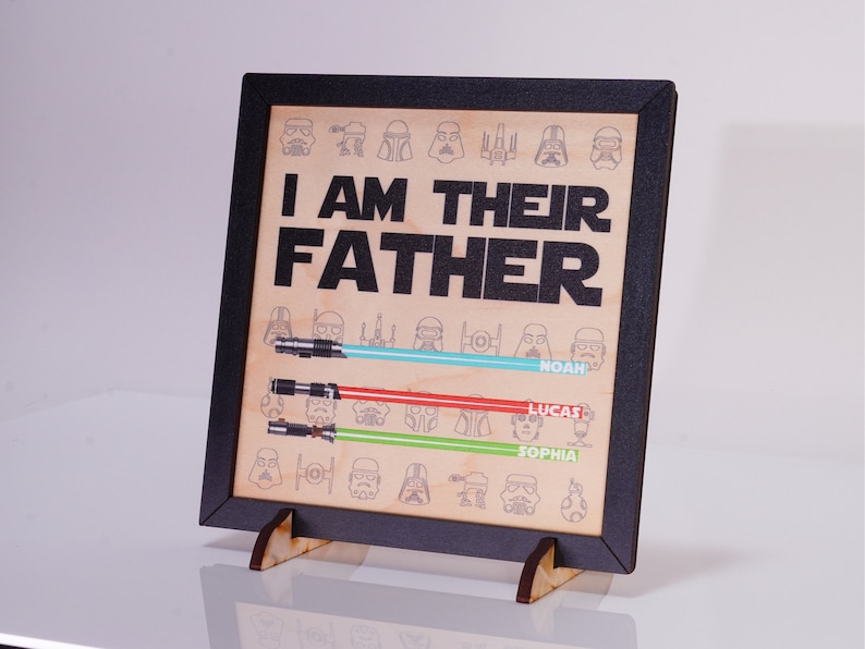 May include: Wooden framed sign with the text "I AM THEIR FATHER" and lightsaber illustrations. The sign includes the names Noah, Lucas, and Sophia. The sign is supported by a wooden stand.